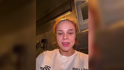 Media: Video of a young woman with light skin and shoulder-length brown hair, smiling, wearing a white T-shirt. She's indoors with dim lighting, standing in front of a dark, textured wall with a window and exposed pipes in the background.