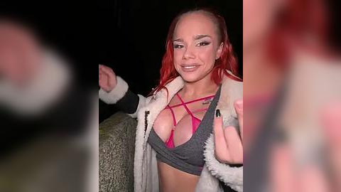 Media: Video of a smiling woman with vibrant red hair, wearing a revealing pink strappy top, gray jacket, and furry collar.