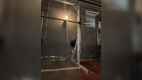 Media: Video of a dimly lit, industrial-style gym with a heavy-duty squat rack, concrete walls, and a worn, reddish-brown rubber floor. The background features a metal door partially open, revealing a dimly lit room beyond.