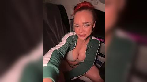 Media: Video of a fair-skinned woman with red hair, wearing a green and white track jacket, revealing a light gray bra, seated in a car, with blurred background.