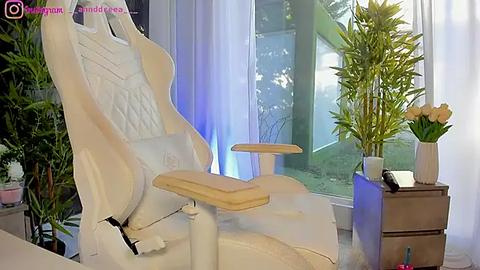 Media: Video of a white gaming chair with a wooden armrest, positioned in front of a large window with bamboo plants and a vase of yellow flowers.