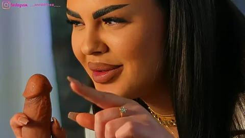 Media: Video of a woman with long black hair, tan skin, and dark eyebrows, holding a large erect penis. She wears a gold necklace and has manicured nails. Background is blurry.