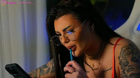 Media: Video of a tattooed woman with long dark hair, wearing glasses, red top, and gold necklace, typing on a smartphone, in a dimly lit room with white curtains.