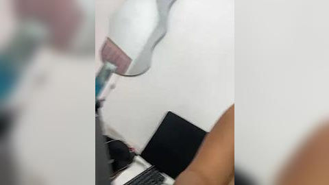 Media: A video shows a close-up of a white desk lamp with a pink shade, partially obscuring a black laptop and other unidentified objects on a white surface. The image is slightly blurry, making the details difficult to discern clearly.