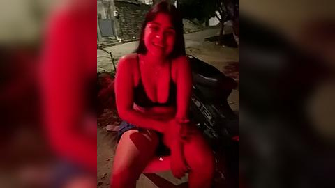 Media: Video of a young woman with medium-length brown hair, wearing a black bra and denim shorts, sitting on a street in dim lighting. Her face is partially obscured by red light.