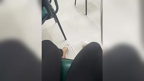 Media: Video of a person's legs in black leggings, feet in white socks, sitting on a green chair in a tiled room.