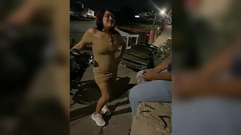 Media: Video of a joyful, curvy Latina woman with medium-length dark hair, wearing a tight, off-the-shoulder beige dress and white sneakers, riding a motorcycle at night in a suburban neighborhood with parked cars and streetlights.