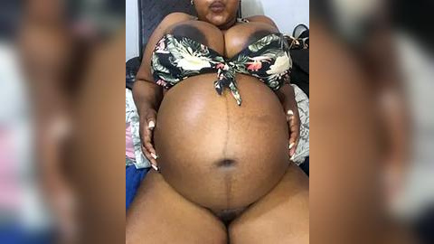 Media: Video of a pregnant Black woman with a large belly, wearing a floral bikini top, seated on a bed. Her skin is dark brown, and her breasts are large. The background is blurry.