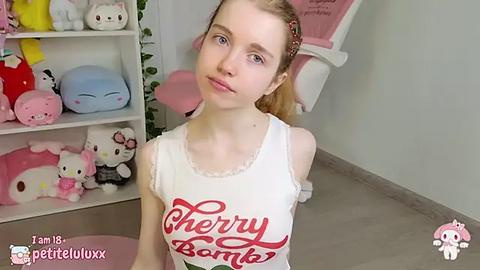 Media: Video of a young, fair-skinned girl with blonde hair, wearing a white tank top with \"Cherry Bomb\" text, kneeling in a pink-themed room filled with Hello Kitty decor.