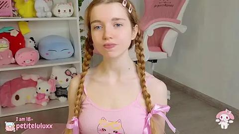 Media: A young, fair-skinned girl with blonde pigtails, wearing a pink tank top with a Hello Kitty design, stands in a Hello Kitty-themed room with plush toys and a pink gaming chair.