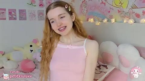 Media: A video of a slender, fair-skinned young woman with long blonde hair in a pink tank top, smiling in a soft, pastel-colored room adorned with plush toys and anime posters.