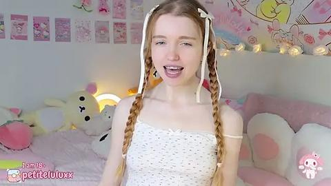 Media: Video of a young, slender, fair-skinned girl with long, blonde braided pigtails, wearing a white knitted dress, smiling in a cozy, pink-themed bedroom.