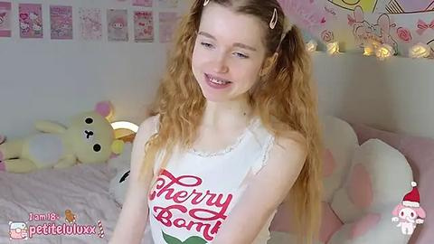 Media: Video of a young, fair-skinned, curly-haired girl in a \"Cherry Bomb\" tank top, smiling in a cozy, pink-themed bedroom with plush toys and fairy lights.