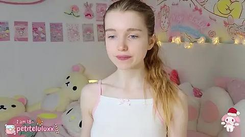 Media: Video of a young, fair-skinned girl with light brown hair in a ponytail, wearing a white tank top, standing in a pastel-themed room filled with plush toys and pink decorations.