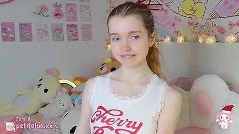 Media: A video of a young, fair-skinned woman with light brown hair, wearing a white tank top with red \"Merry\" text, sitting amidst plush toys in a festive room.