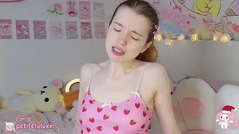 Media: Video of a young, fair-skinned woman with straight blonde hair in a pink strawberry-patterned camisole, smiling, surrounded by plush toys and a soft, pink-themed bedroom.