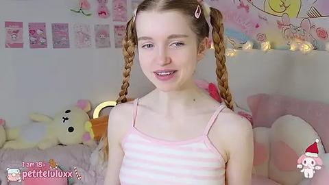 Media: Video of a young, slender, fair-skinned girl with braided hair in a pink striped tank top, smiling in a pink-themed room with plush toys and wall art.