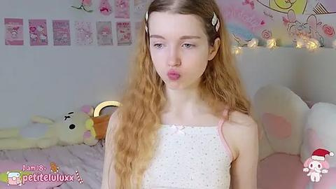 Media: A video of a young, fair-skinned girl with long, wavy blonde hair, wearing a white polka-dot nightgown, blowing a kiss, against a pink, cartoon-themed bedroom backdrop.