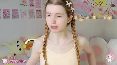 Media: Video of a young, fair-skinned woman with long, blonde braided hair adorned with bows, wearing a yellow tank top. Background features pastel-colored wall decorations, stuffed animals, and a cozy atmosphere.