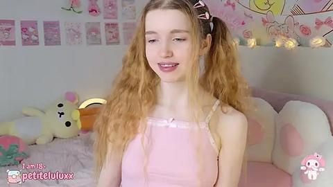 Media: Video of a young, slim, fair-skinned girl with long, curly blonde hair in pigtails, wearing a light pink spaghetti strap dress, sitting in a pastel-themed bedroom with plush toys and fairy lights.