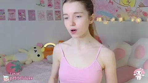 Media: A video of a pale-skinned, slim young woman with long blonde hair wearing a pink spaghetti-strap top. She's in a pink, cartoon-themed bedroom with plush toys and pastel decorations.