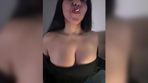 Media: A close-up video of a woman with long black hair and medium skin tone, wearing a black off-the-shoulder top, slightly puckered lips, and visible cleavage.