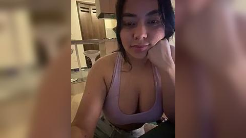 Media: Video of a young woman with medium skin tone, dark hair, and large breasts, wearing a light purple sports bra, leaning on a countertop in a dimly lit kitchen.