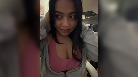 Media: Video of a young woman with medium-dark skin and long black hair, wearing a pink tank top and unzipped denim jacket, in a dimly lit kitchen with blurred background.