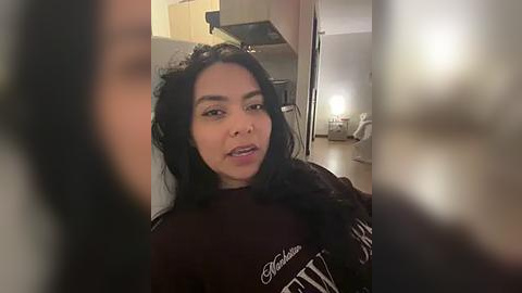 Media: Video of a woman with long black hair, wearing a dark sweater, standing in a dimly lit kitchen. The background features a stove and cabinets.