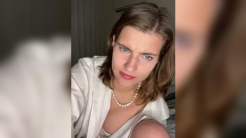 Media: Video of a light-skinned woman with shoulder-length brown hair, wearing a white robe and pearl necklace, looking angry and serious in a dimly lit room.