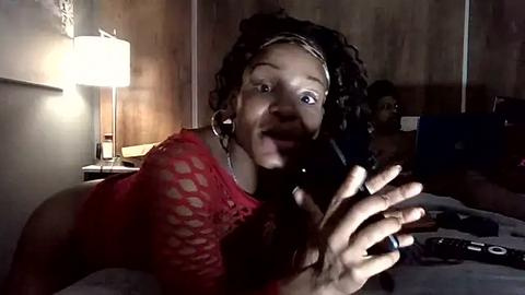 Media: A dimly-lit video of a young African-American woman with short dreadlocks, wearing a red fishnet top, holding a phone, smiling, in a dimly-lit room with a table lamp and laptop.