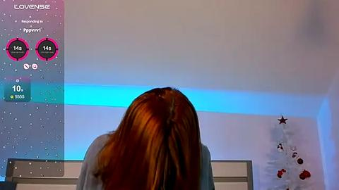 Media: Video of a woman with long red hair facing away, in a room with a blue light, a digital clock, and a decorative star garland.