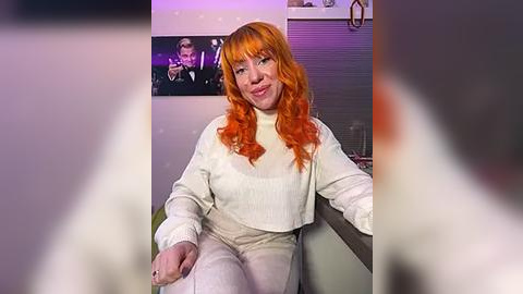 Media: A video of a smiling woman with vibrant orange hair and fair skin, wearing a white turtleneck and pants, sitting on a counter. Background includes a framed photo of a man and a metallic kitchen backsplash.