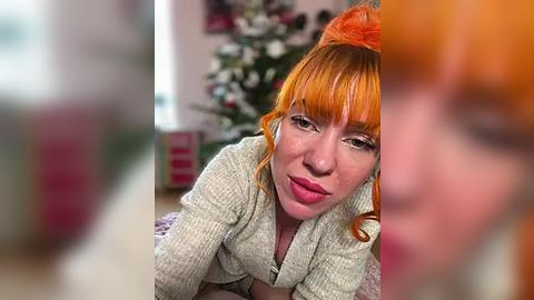 Media: Video of a fair-skinned woman with bright orange hair and bangs, wearing a beige cardigan, kneeling on a carpeted floor. Background features a blurred Christmas tree and decor.