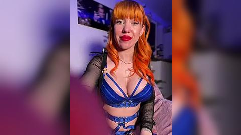 Media: Video of a fair-skinned woman with orange hair and blue lingerie, smiling, in a dimly lit room with blurred figures in the background.