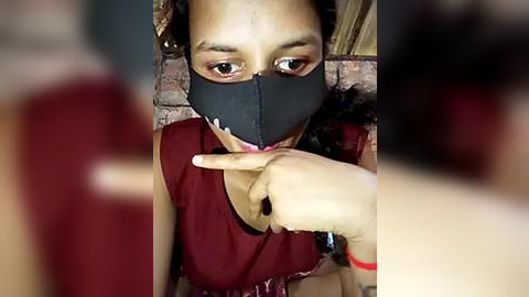 Media: Video of a woman with medium brown skin and medium-length black hair, wearing a black face mask, red top, and red bracelet, covering her mouth with her index finger. Background shows blurred brick wall.