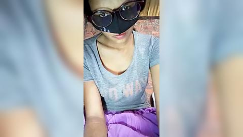 Media: Video of a young Asian woman with glasses and a face mask, wearing a gray T-shirt with \"Nurse\" printed on it, seated against a brick wall, legs in a purple skirt.