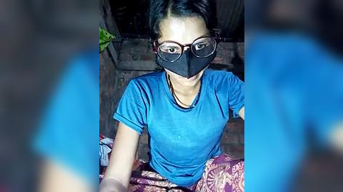 Media: Video of an Asian woman with short black hair, wearing a blue t-shirt, black face mask, and glasses, seated on a patterned fabric, indoors with brick walls and green plant in background.