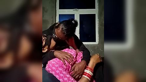 Media: Video of an intimate moment between two women in a dimly lit room. One woman with dark skin and braids embraces the other, who is wearing a pink dress with white patterns and multiple red bangles. The background shows a window with dark curtains.