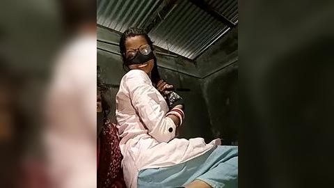 Media: Video of a woman with dark skin and glasses, wearing a pink hospital gown and white gloves, sitting in a dimly lit room with corrugated metal walls and a red blanket.