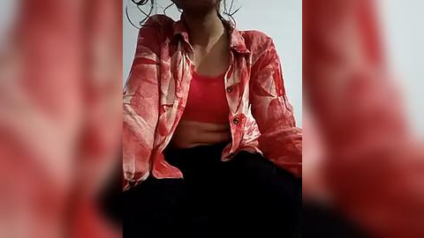 Media: Video of a woman with medium skin tone, wearing a red-patterned blouse over a red bra, and black pants, taken from an upward angle, with blurred hands in the foreground.