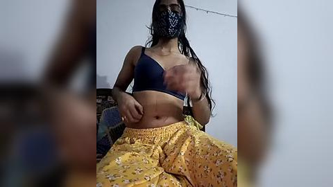 Media: A video of a young South Asian woman with long black hair, wearing a blue bra, yellow floral skirt, and black face mask, sitting on a bed, slightly blurred.