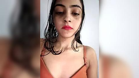 Media: Video of a young woman with wet, dark hair, wearing a strapless, orange top, and red lipstick, standing in a blurred, plain white background.