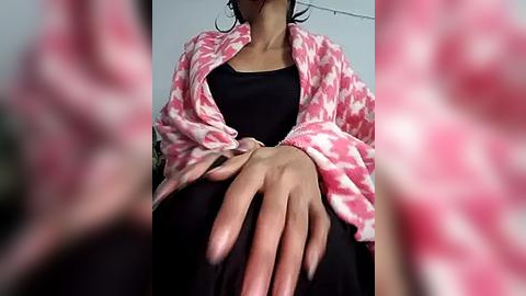 Media: Video of a woman in a black tank top, wearing a pink and white patterned robe, with her hands clasped in front, indoors.