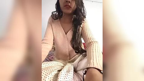 Media: Video of a young woman with long, wavy dark hair, wearing a ribbed, beige, V-neck sweater and white, patterned pants, seated indoors with a soft-focus background.