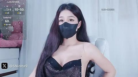 Media: A video of an Asian woman with long black hair, wearing a black face mask, a black sequined dress, and a black bra, sitting against a white curtain.