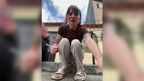 Media: Video of a tattooed, dark-haired woman in a brown top and beige pants squatting on a ledge, holding a cigarette, with a blurred background of a street and building.