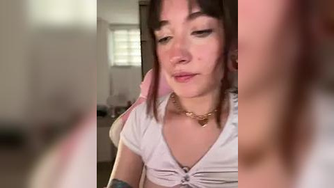 Media: Video of a young woman with fair skin, straight brown hair, and bangs, wearing a white crop top, seated indoors. Background includes blurred kitchen elements.