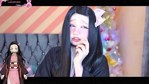 Media: Video of a cosplay: a woman with long black hair, wearing a black kimono and pink makeup, holding her cheek. Background features a Christmas tree and striped wall.