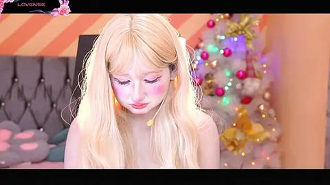 Media: Video of a young, pale-skinned woman with long blonde hair, wearing blush and earrings, sitting on a bed in a colorful, festive room with a decorated Christmas tree.